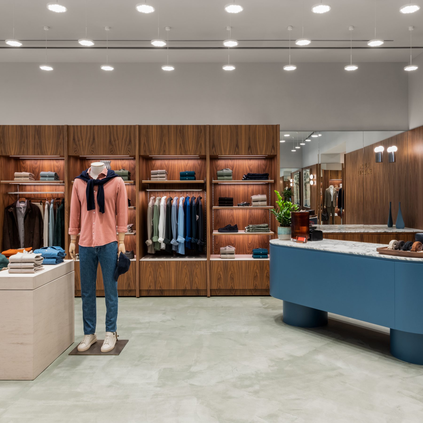 a modern menswear store featuring warm wood-paneled walls, a curved blue checkout counter, mannequins displaying casual outfits, and a seated lounge area